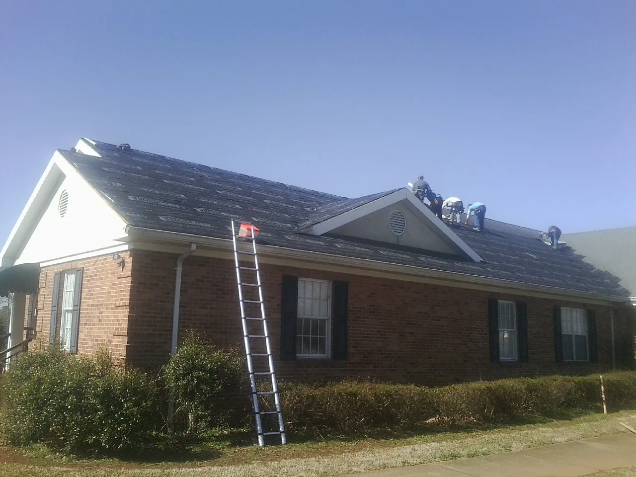 Crew tearing shingles off a brick ranch roof with a ladder set — Emergency Roof Repair work by our crew in New Johnsonville