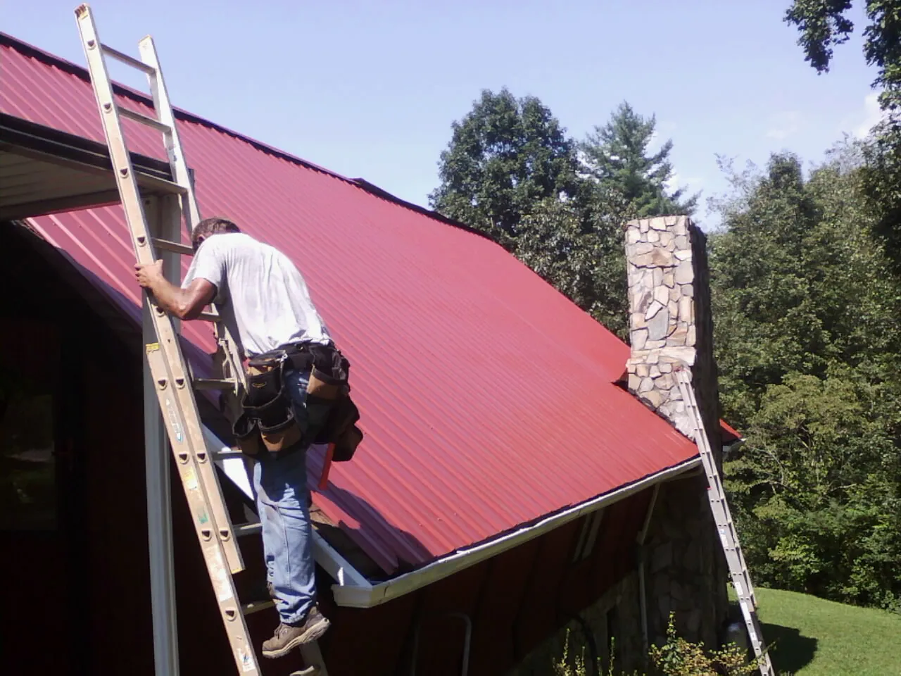 Roofer on a ladder beside a red metal roof and stone chimney — Soffit & Fascia Repair by Roofing Brothers in New Johnsonville